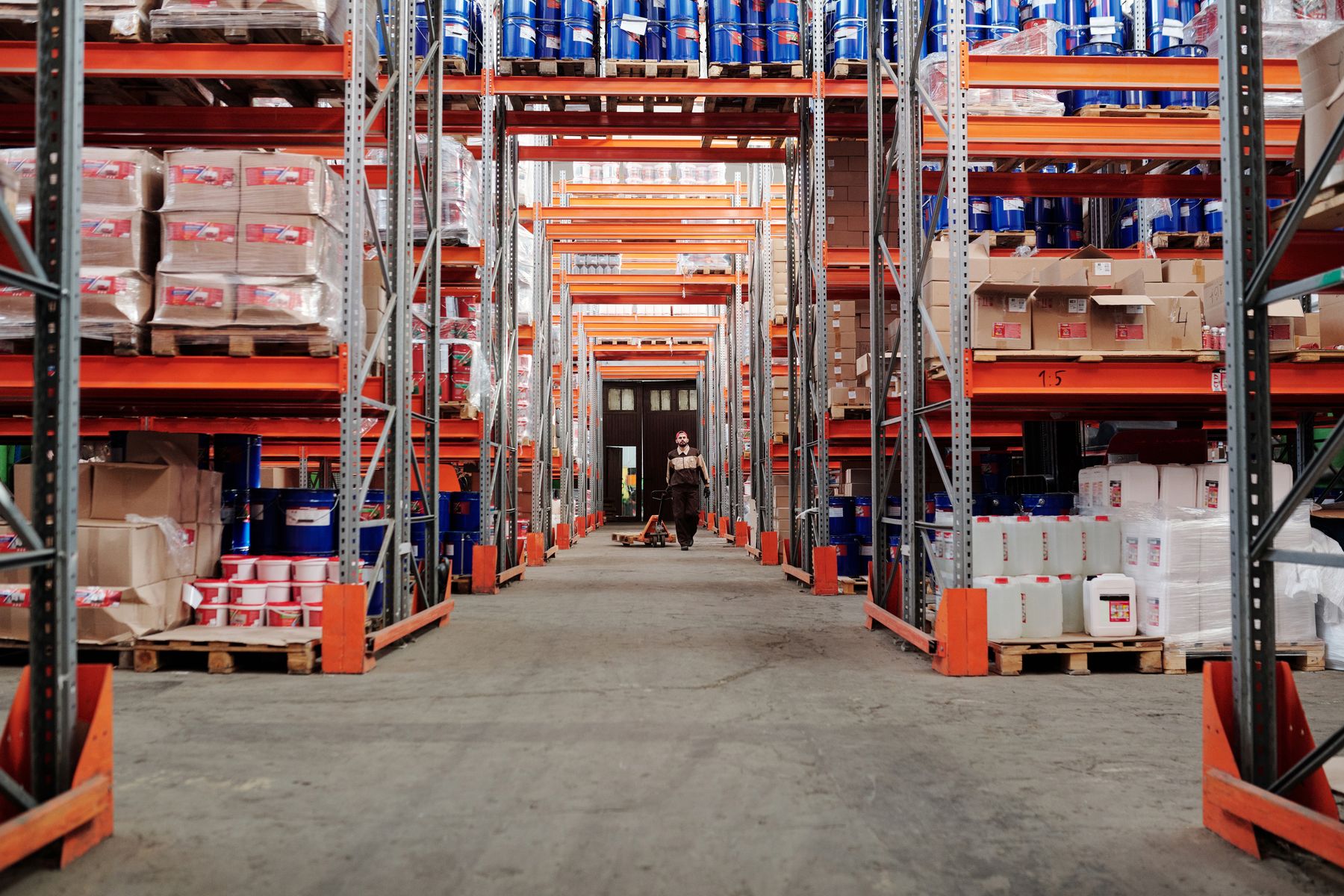 Warehouse aisle lined with stocked pallet racks and a worker in the distance